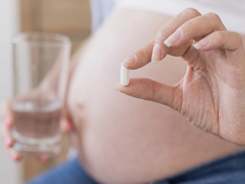 Close-up da mão de uma mulher grávida segurando um comprimido branco de cálcio, com a barriga de gravidez e um copo de água visíveis ao fundo.