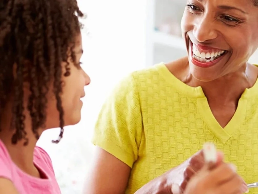 Uma mulher idosa sorridente e uma menina com cabelo cacheado riem juntas à mesa de uma cozinha bem iluminada. Elas compartilham um momento feliz, inspirando o cuidado com a saúde através de uma alimentação rica em cálcio no café da manhã.