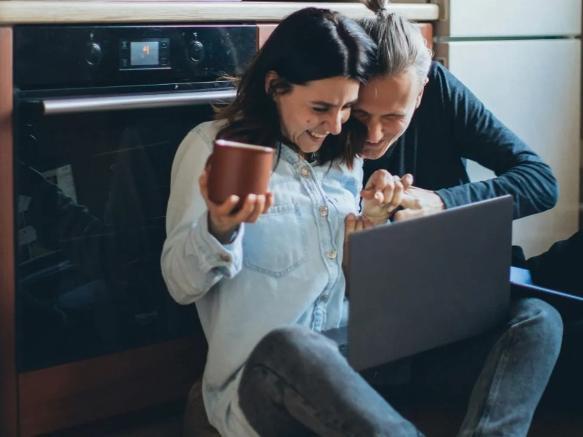 Um casal feliz usa um notebook na cozinha, representando a vida a dois e como a dupla de nutrientes vitamina D e cálcio são importantes para a saúde óssea.