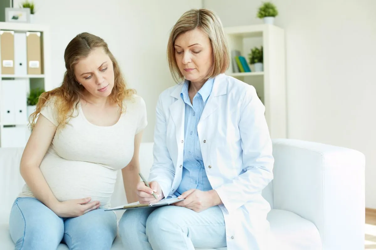 Médica de jaleco branco anota recomendações em uma prancheta para uma mulher grávida sentada ao seu lado em um sofá, que a observa atentamente.