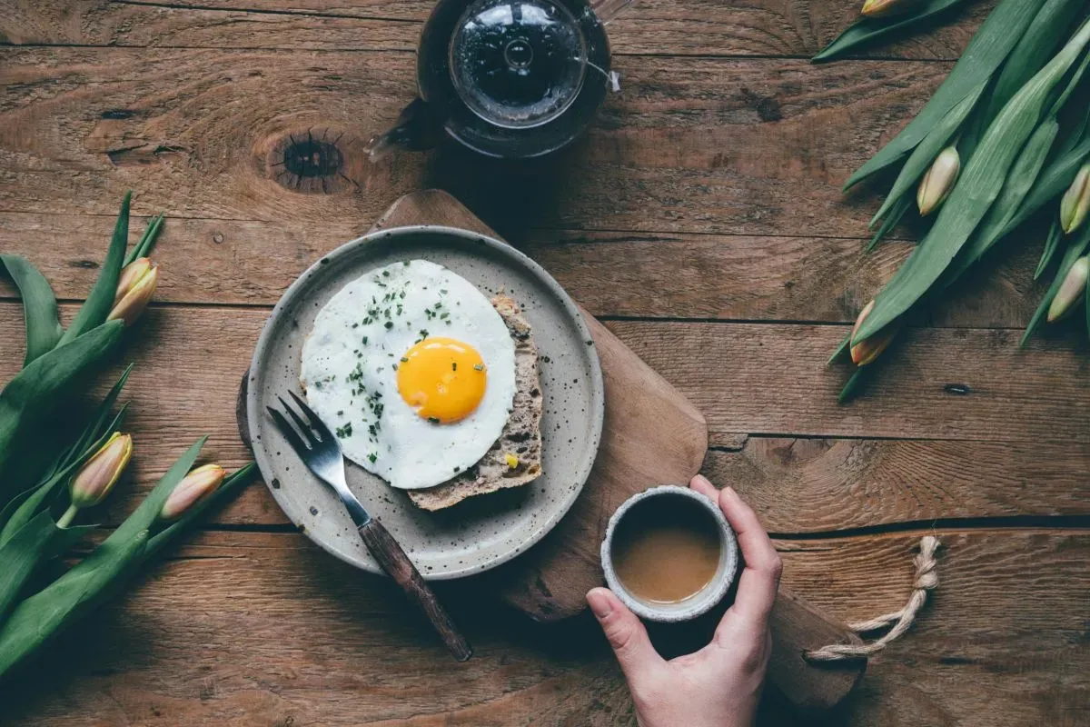 A imagem de um ovo frito sobre uma torrada, acompanhado de uma xícara de café, ilustra a ingestão da vitamina D junto ao café da manhã, pois a presença de gordura nos ovos melhora sua absorção.