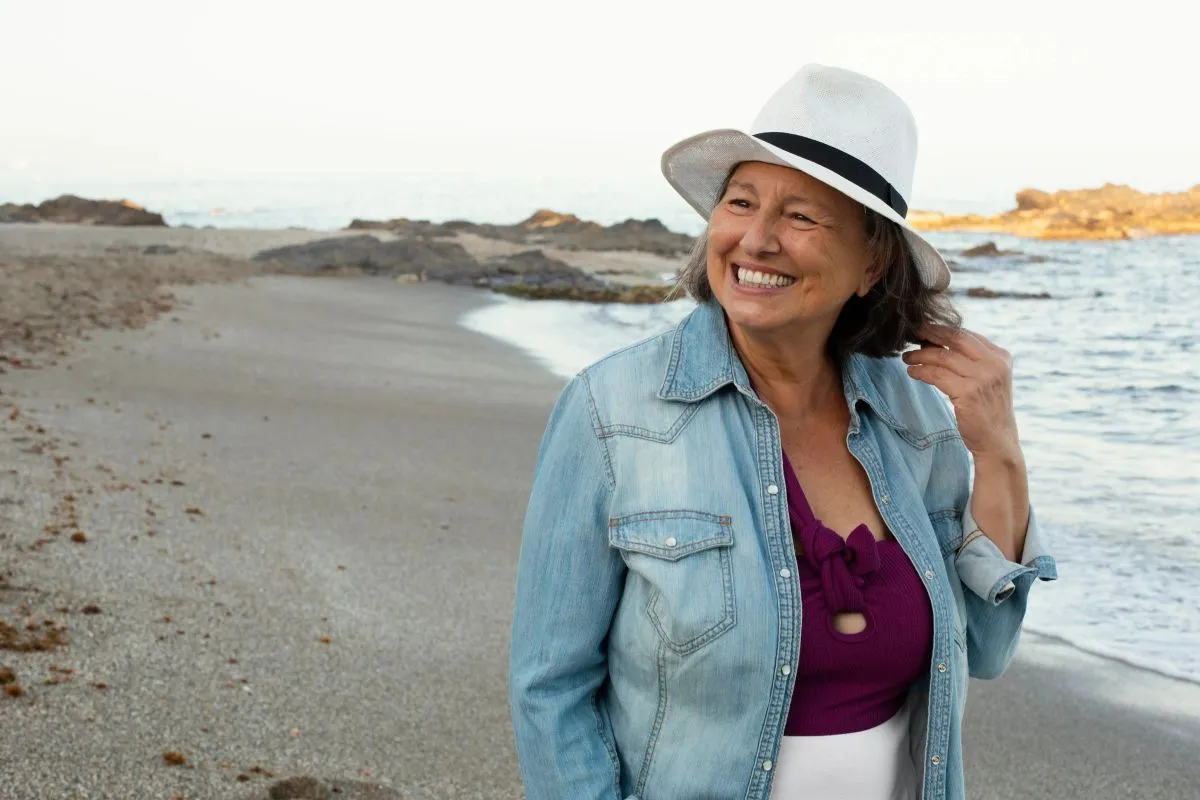 Uma mulher mais velha, sorrindo e com aparência saudável, usa um chapéu branco em uma praia, representando os benefícios dos suplementos para idosos na manutenção da vitalidade.
