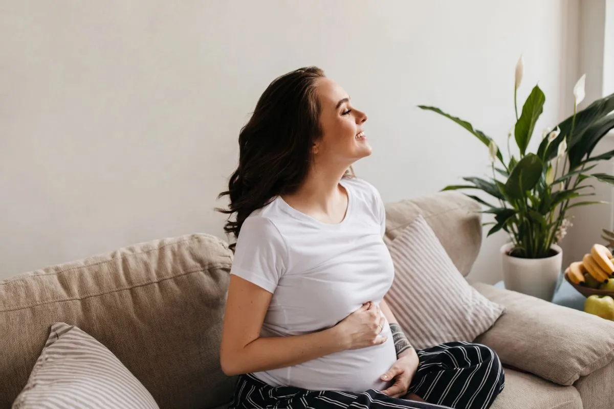 Mulher grávida sorrindo de olhos fechados, sentada em um sofá, com as duas mãos sobre a barriga, transmitindo uma sensação de saúde e tranquilidade na gestação.