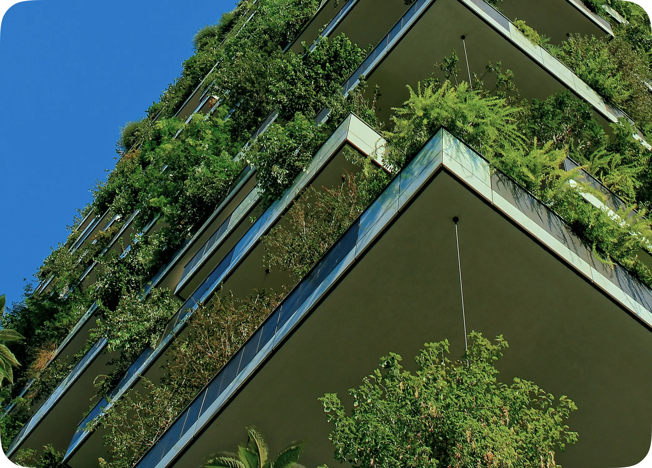 Fachada de um edifício moderno visto de baixo para cima, com as varandas repletas de plantas e árvores sob um céu azul.