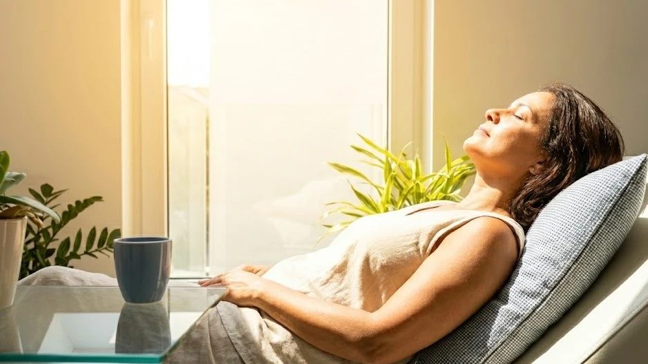 Uma mulher de meia-idade reclina confortavelmente com os olhos fechados sob forte luz natural do sol que entra por uma janela, banhando seu rosto e braços. Ela está em um ambiente moderno e clean, com uma manta com toque azul sutil no sofá. 