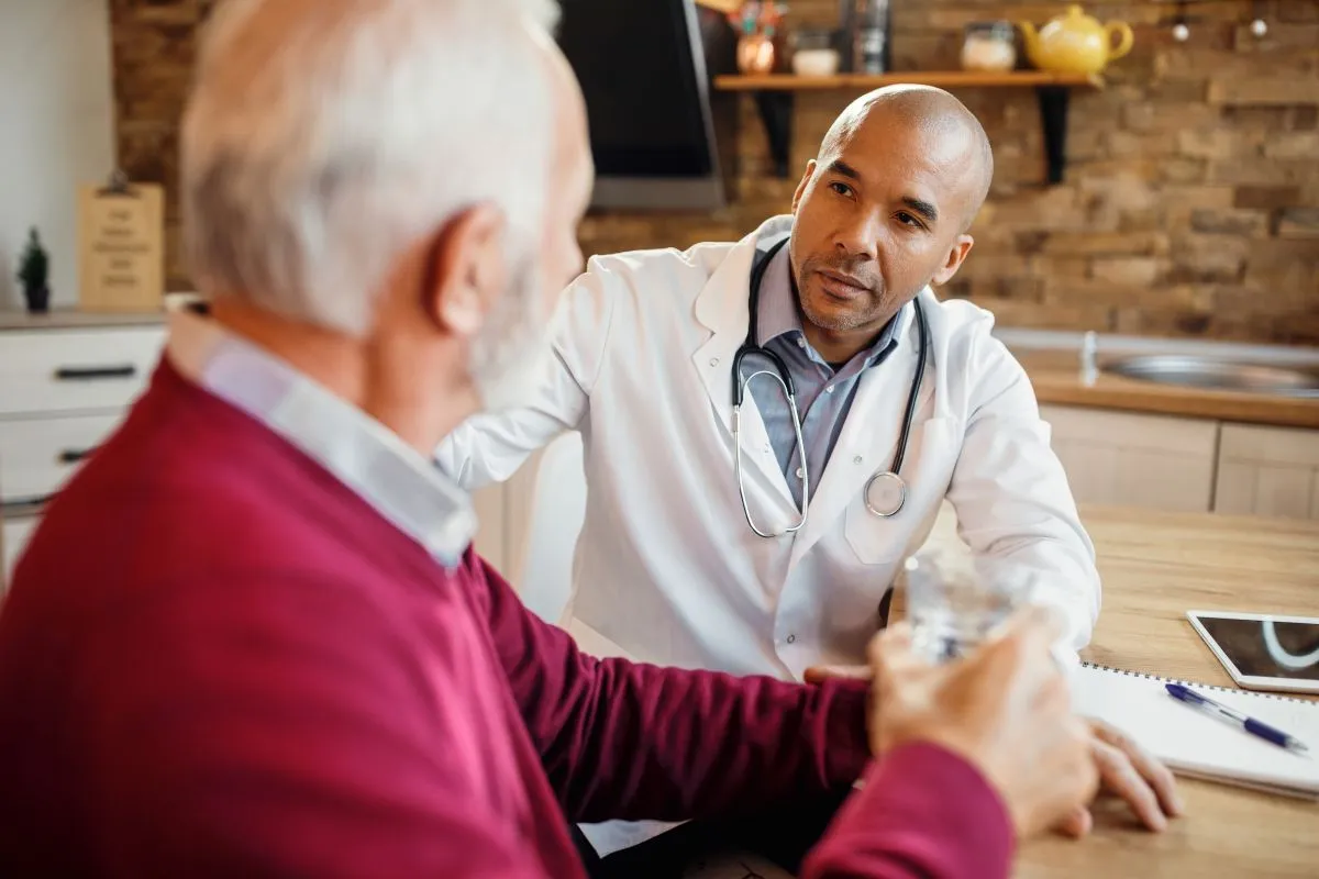 Um médico atencioso, vestindo jaleco branco e estetoscópio, conversa com um paciente idoso em um ambiente que parece ser uma casa ou consultório informal. O paciente segura um copo d'água, e a interação sugere uma discussão sobre a condição de saúde, como a hipocalcemia.